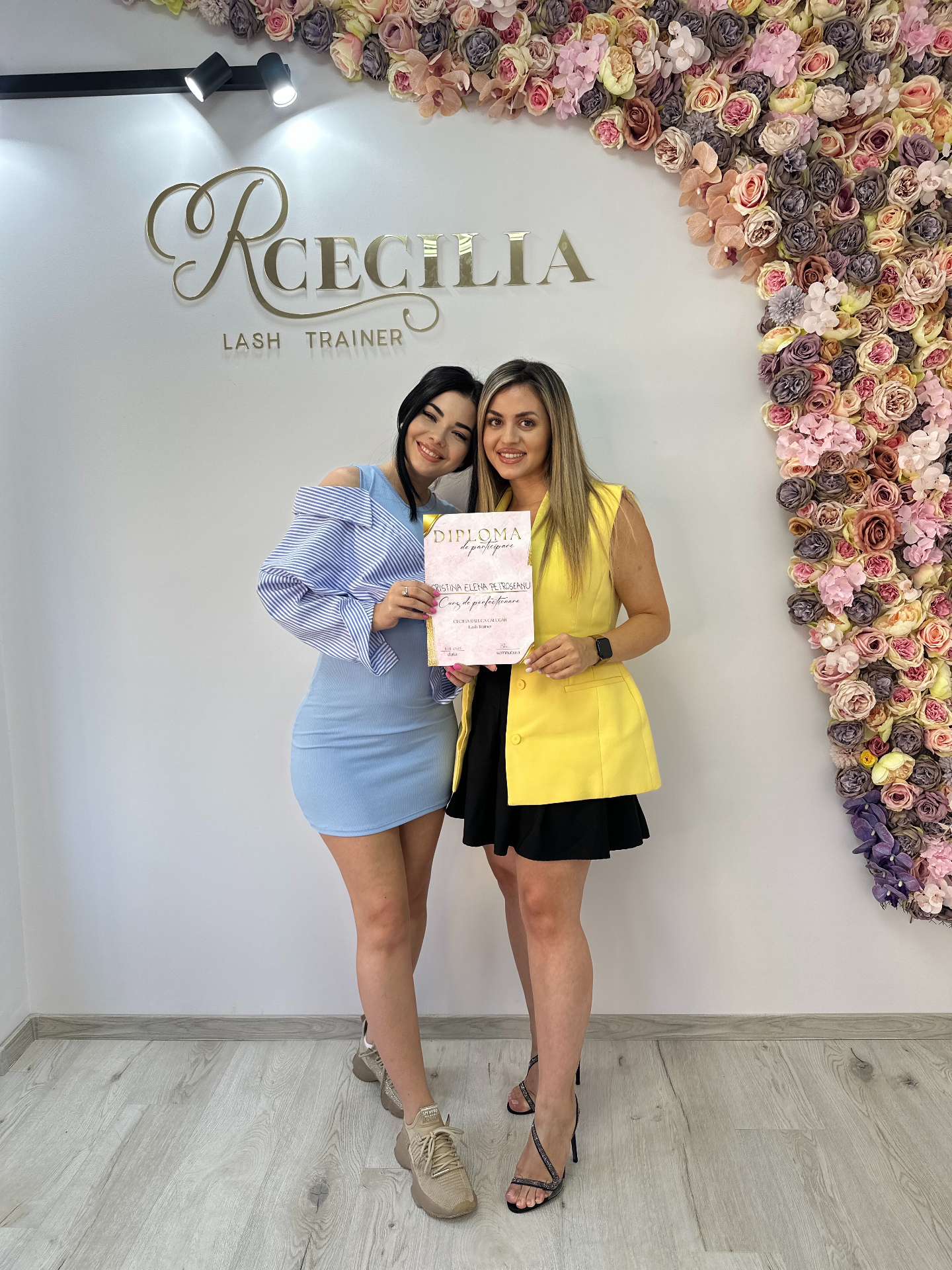 Cursantă VIP Private Training cu diploma de perfecționare, în fața peretelui floral din academia Cecilia Călugăr Timișoara